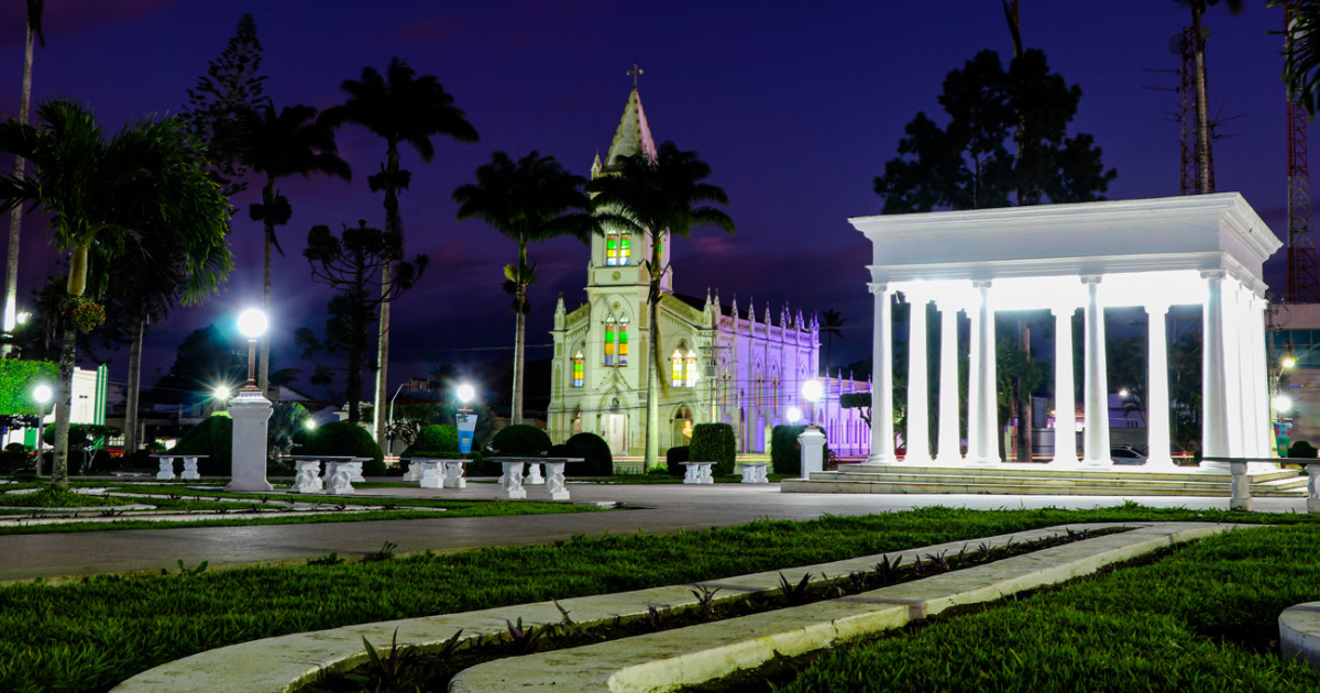 A imagem é uma foto noturna de uma praça com uma igreja iluminada ao fundo e uma estrutura com colunas brancas à direita. A igreja tem uma arquitetura gótica com vitrais coloridos.